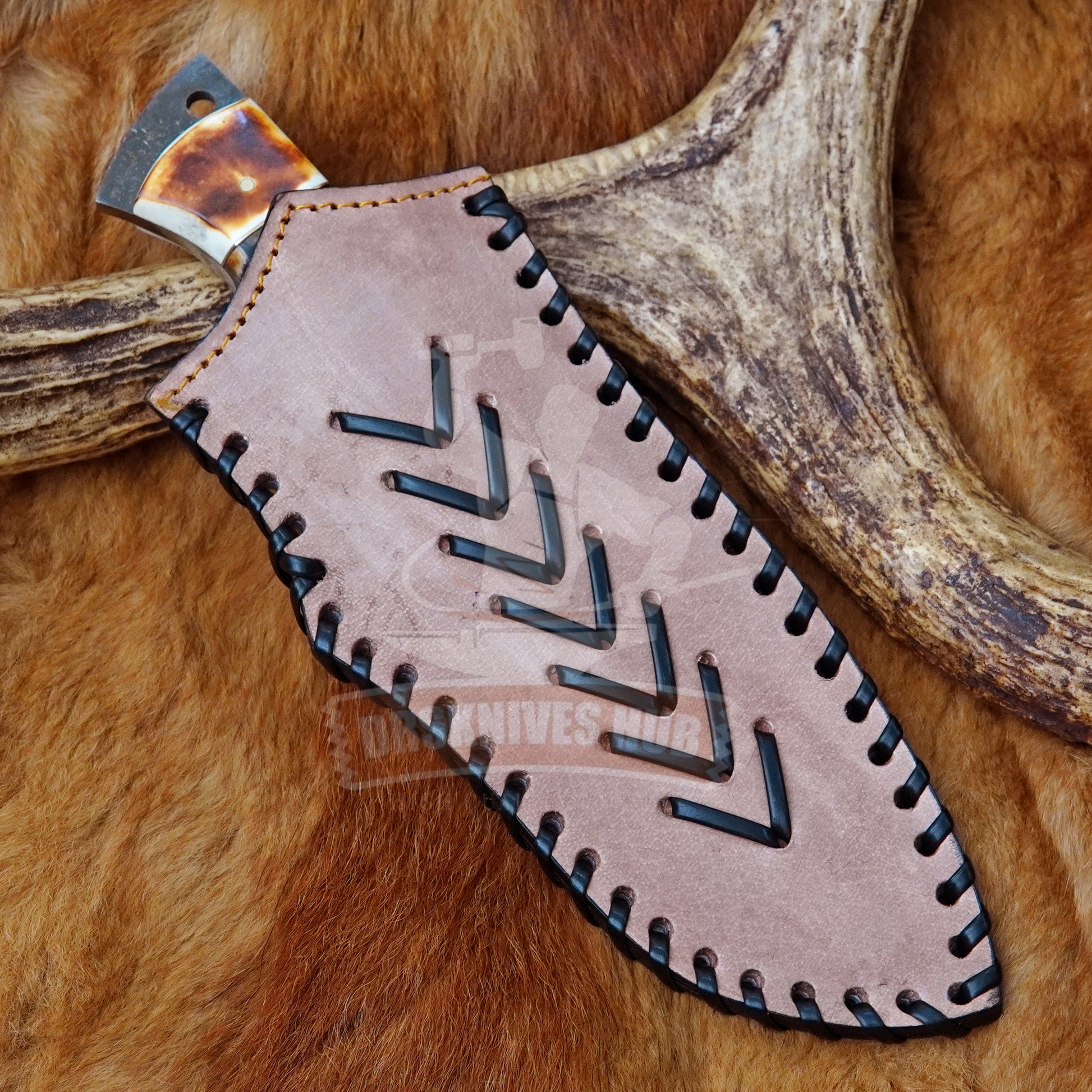 A close-up of a knife partially tucked into a tan leather sheath with black stitched edges and a cutout chevron pattern, placed on brown fur with antlers in the background.