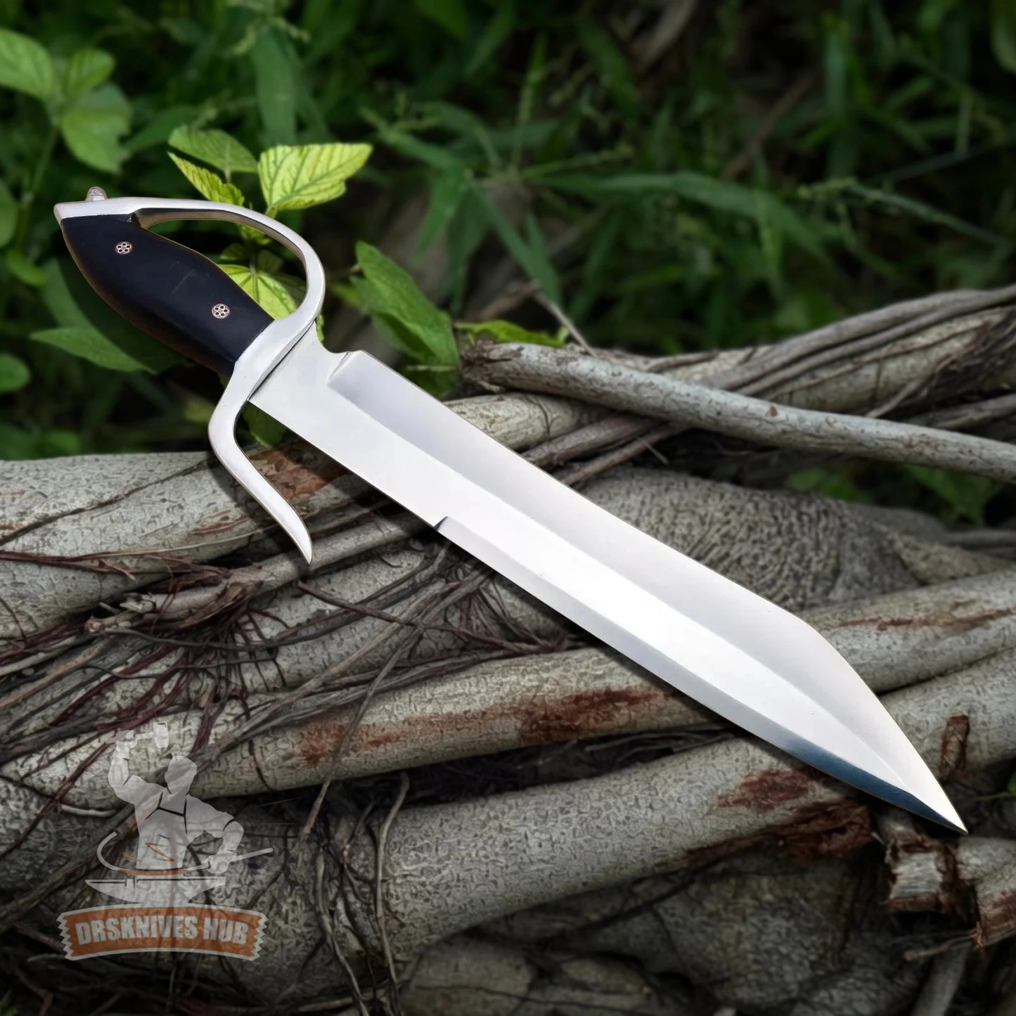 A fixed-blade knife with a polished silver blade, curved guard, and black handle with brass rivets, resting on a pile of tree roots and branches outdoors with green foliage in the background.