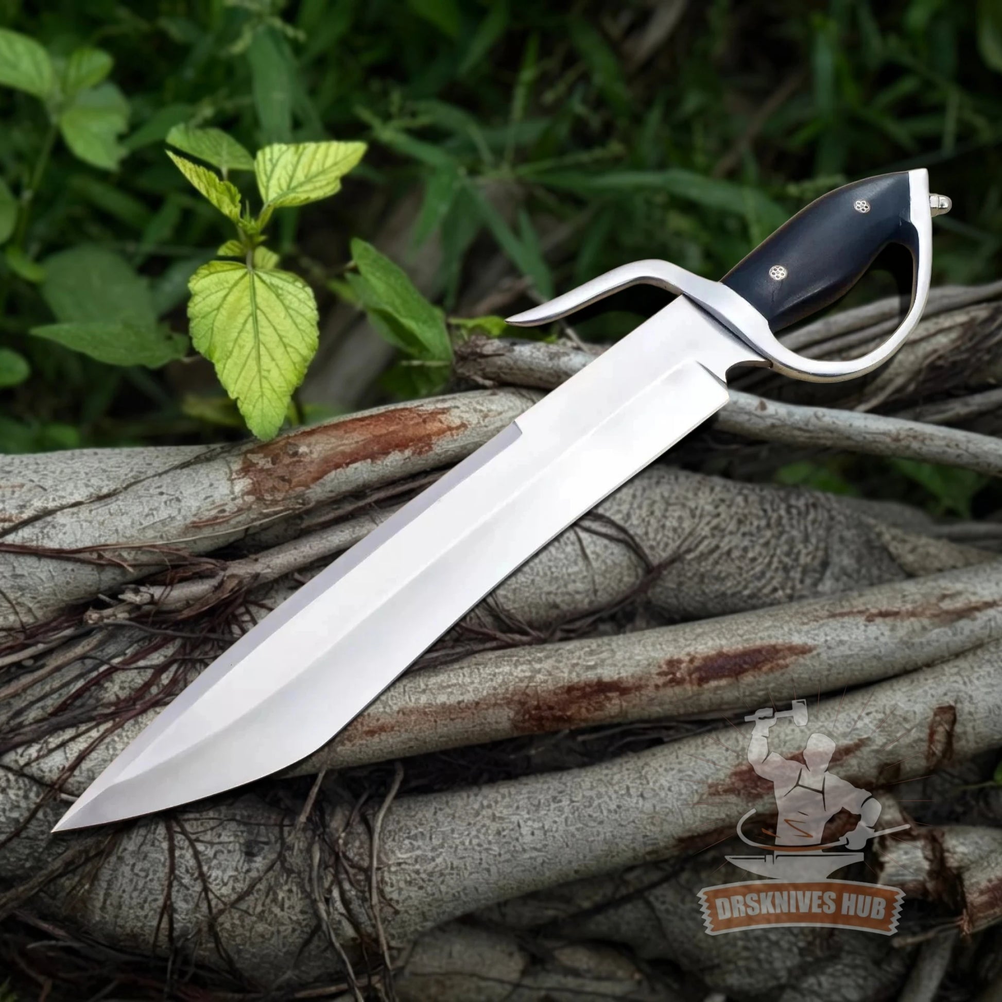 A large fixed-blade knife with a polished silver blade and black handle, resting diagonally on a pile of tree roots with green foliage in the background.