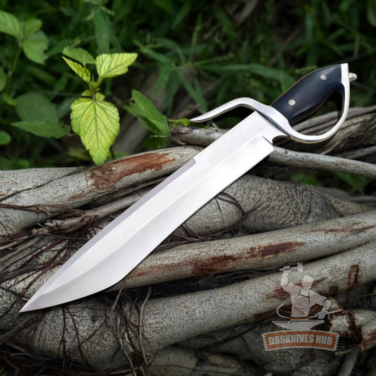 A large fixed-blade knife with a polished silver blade and black handle, resting diagonally on a pile of tree roots with green foliage in the background.