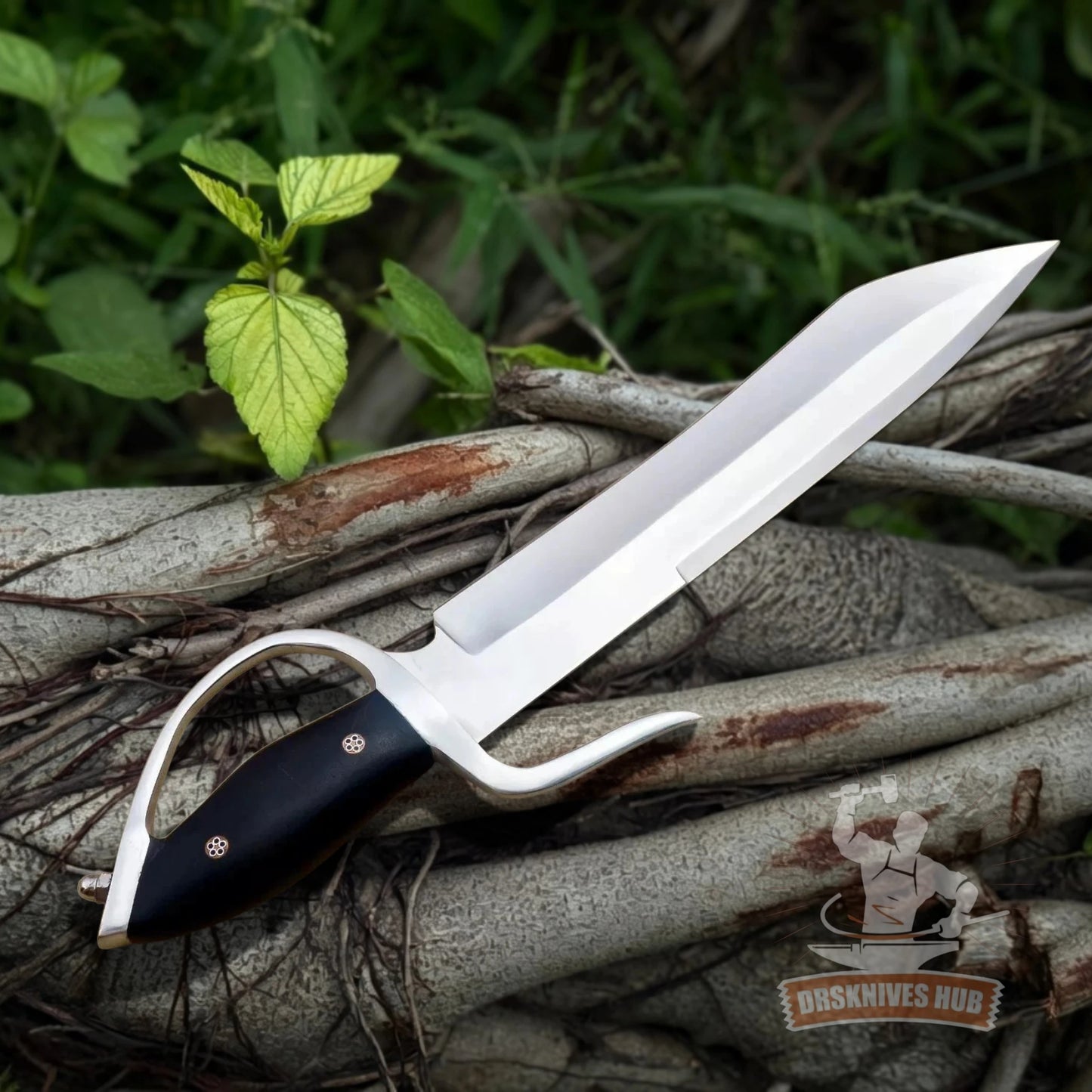 A fixed-blade knife with a silver blade and black handle, resting on a pile of tree roots outdoors with green foliage in the background. A 'Dusknives Hub' logo is visible in the bottom right corner.