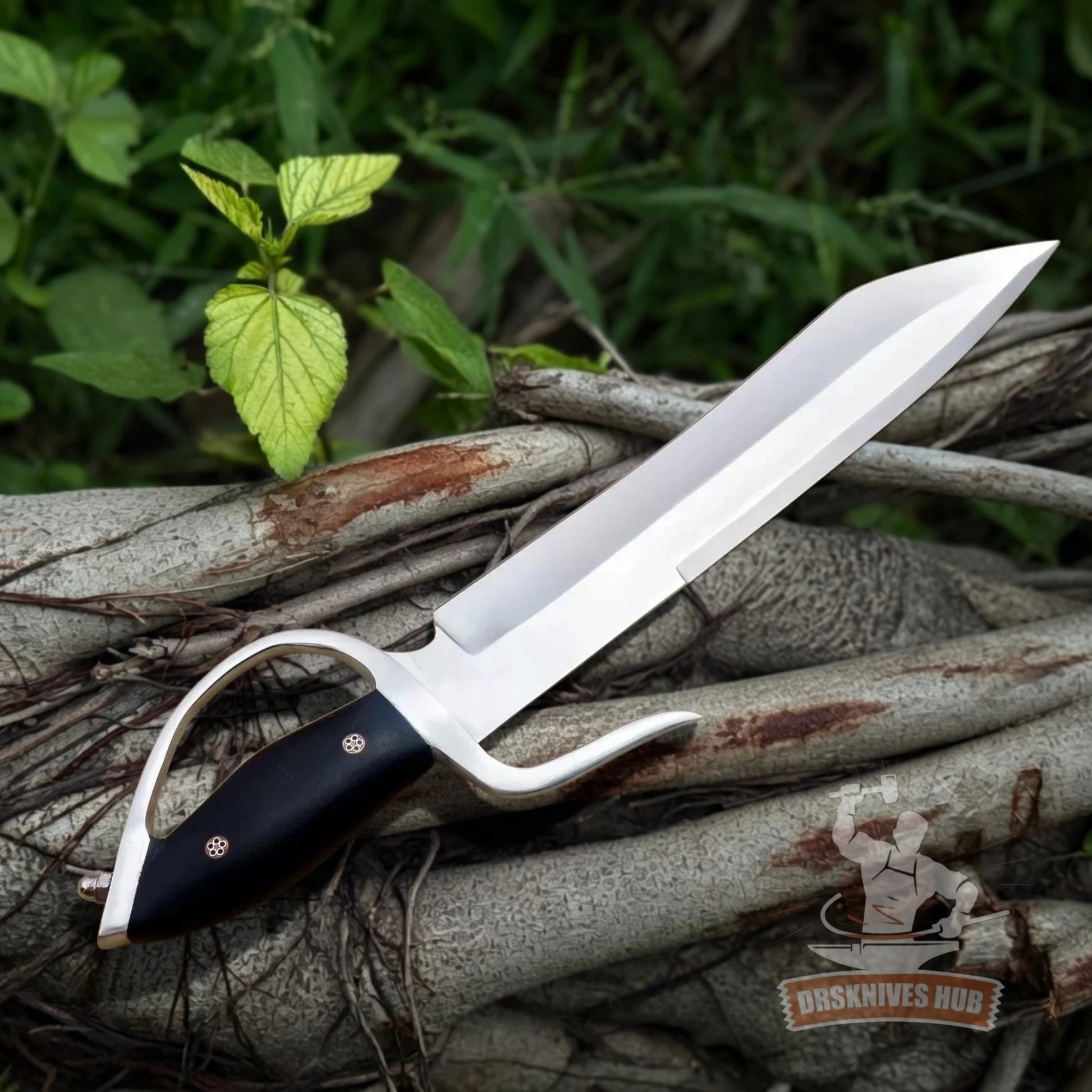 A fixed-blade knife with a silver blade and black handle, resting on a pile of tree roots outdoors with green foliage in the background. A 'Dusknives Hub' logo is visible in the bottom right corner.