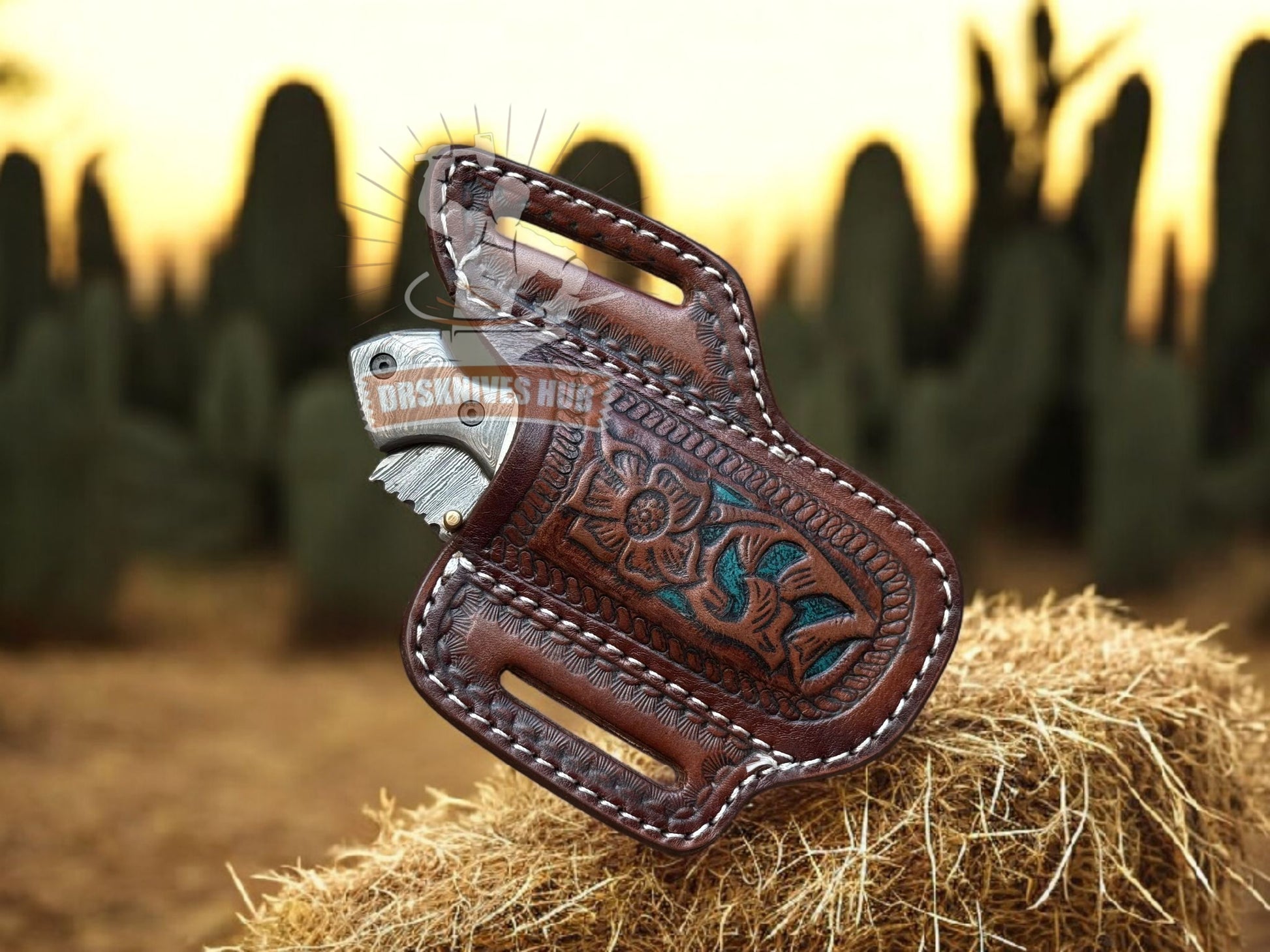 A brown tooled leather knife sheath holding a folding knife with a silver blade, displayed on a bale of hay with a blurred outdoor desert background.